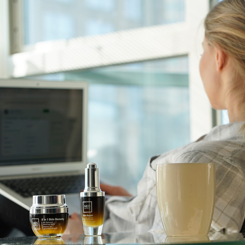 Hormone-freindly beauty rituals by NeoNaturelle. 5-in-1 Skin Remedy and Skin Repair Serum, on a glass table with a coffee cup and a woman working on a laptop in the background.